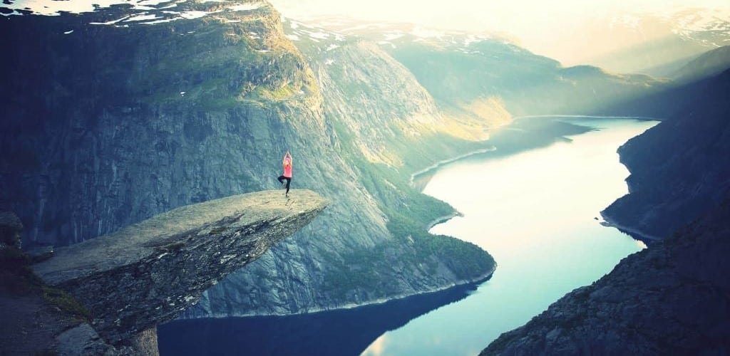 Photo of woman in a yoga tree pose, on top of a rocky ledge, surrounded by mountainous scenery, with a lake or river far below her.