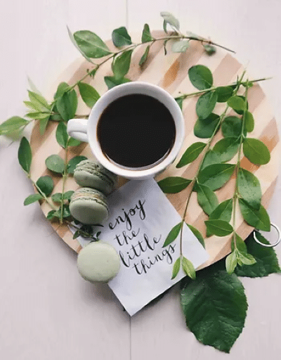 Birds eye image of a cup of coffee on a wooden board, next to some foliage, with green macarons, and a note saying 'enjoy the little things'.