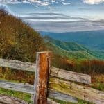 Outdoor scene of rolling green hills, with low cloud in the far distance, and a wooden fence in the foreground.